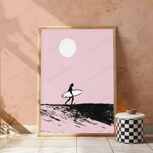 May include: A framed wall art print featuring a black silhouette of a person carrying a surfboard across a textured black landscape against a solid light pink background with a white circular sun positioned in the upper center of the composition.