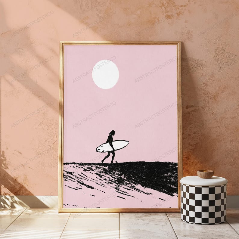 May include: A framed wall art print featuring a black silhouette of a person carrying a surfboard across a textured black landscape against a solid light pink background with a white circular sun positioned in the upper center of the composition.