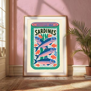 May include: A framed wall art print featuring a stylized illustration of a tin of sardines. The design displays three blue and pink fish stacked vertically, surrounded by small orange fish and green seaweed, with the text SARDINES at the top.