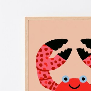 May include: A framed wall art print featuring a stylized crab illustration on a light peach background. The crab has a bright red body, light blue eyes, and pink pincers covered in dark red polka dots with black tips, all encased in a light wood frame.