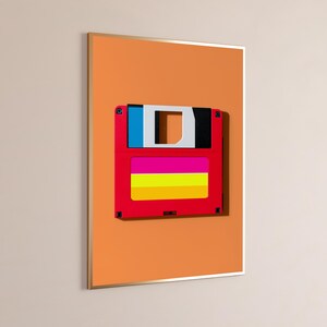 May include: A framed wall art print featuring a red floppy disk with a blue, white, and black top section and a horizontal label with pink, yellow, and orange stripes, set against a solid orange background within a thin metallic frame.