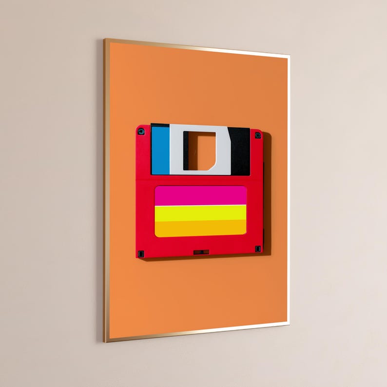 May include: A framed wall art print featuring a red floppy disk with a blue, white, and black top section and a horizontal label with pink, yellow, and orange stripes, set against a solid orange background within a thin metallic frame.