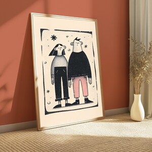 May include: A framed rectangular wall art print featuring a stylised illustration of two figures standing side by side. The figure on the left wears a grey top and dark trousers, while the figure on the right wears a dark top and light pink trousers.