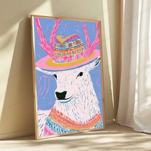 K&ouml;nnte beinhalten: Ein gerahmter Kunstdruck, der einen stilisierten wei&szlig;en Hirsch mit rosa Geweih zeigt, der einen farbenfroh gemusterten Hut und einen passenden Pullover vor einem hellblauen Hintergrund tr&auml;gt. Das Bild nutzt kr&auml;ftige Linien und leuchtende Farben.