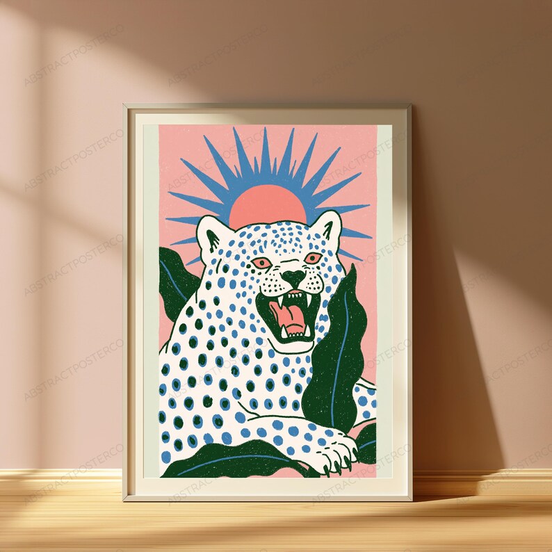 May include: A framed art print featuring a white leopard with blue spots, a roaring expression, and pink eyes. The leopard is surrounded by dark green leaves against a pink background with a blue sunburst design positioned behind its head.