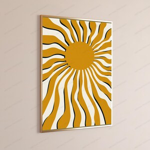 May include: A rectangular wall art print in a thin gold frame featuring a central mustard yellow circle with radiating wavy lines in mustard yellow, cream, and thin black accents, creating a sunburst pattern on a cream-colored background.