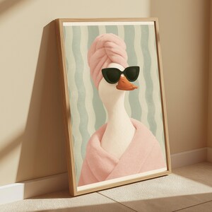 Puede incluir: Una l&aacute;mina de arte mural enmarcada que muestra un pato blanco con una toalla rosa envuelta en la cabeza, un albornoz rosa a juego y gafas de sol negras de ojo de gato. El fondo presenta rayas verticales onduladas en verde claro y blanco.