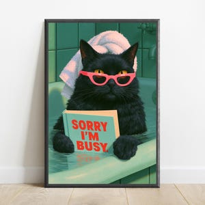 May include: A framed wall art print featuring a black cat in a bathtub, wearing bright pink glasses and a white towel on its head. The cat holds a light blue book with the red text SORRY I'M BUSY printed on the cover, set against a green tiled wall.