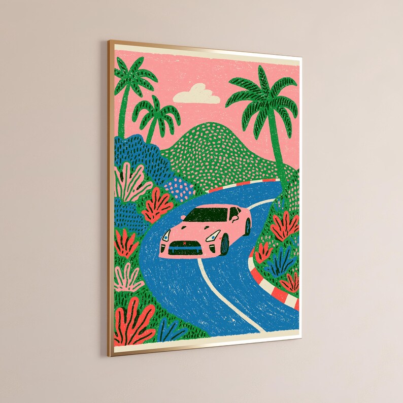 May include: A framed art print featuring a pink sports car driving along a winding blue road. The scene includes green hills, palm trees, and pink foliage against a pink sky with a single white cloud. The road has a red and white striped border.