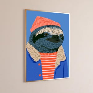 May include: A framed wall art print featuring a stylized illustration of a sloth wearing a coral-colored beanie, a blue jacket with a beige faux-fur collar, and a red and white horizontally striped shirt against a solid blue background.