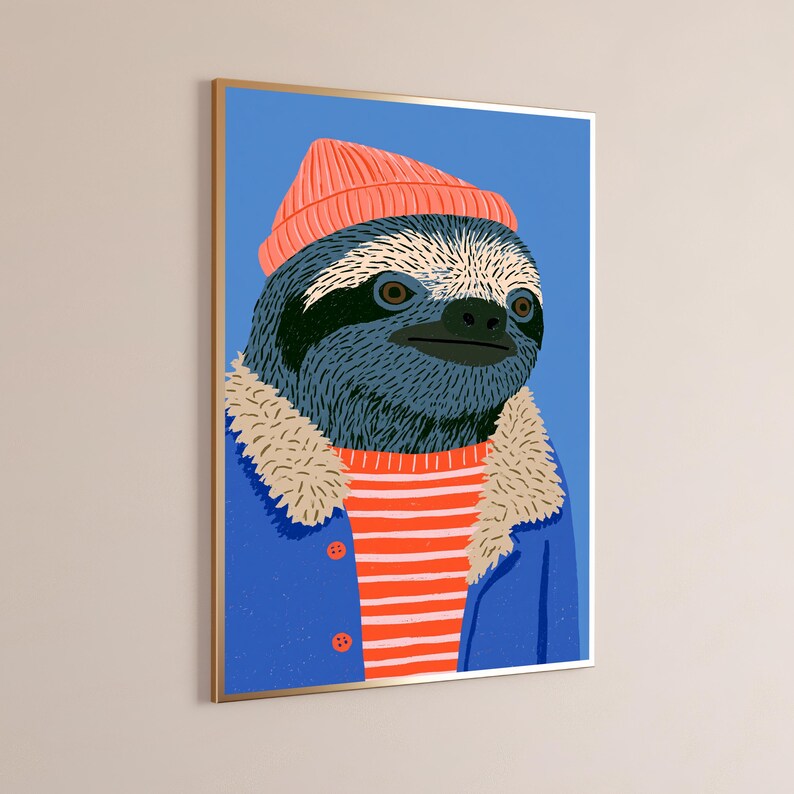 May include: A framed wall art print featuring a stylized illustration of a sloth wearing a coral-colored beanie, a blue jacket with a beige faux-fur collar, and a red and white horizontally striped shirt against a solid blue background.