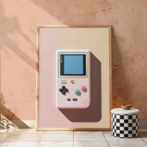 May include: A framed vertical wall art print featuring a light pink handheld video game console with a blue screen, a black directional pad, and red and green circular buttons, set against a soft gradient background of pale pink and cream.