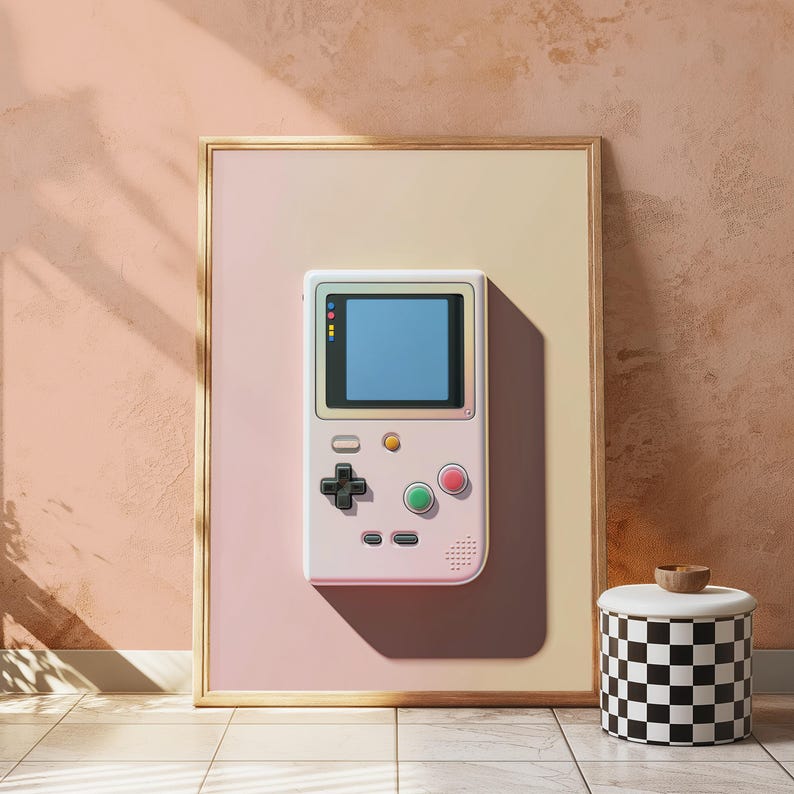 May include: A framed vertical wall art print featuring a light pink handheld video game console with a blue screen, a black directional pad, and red and green circular buttons, set against a soft gradient background of pale pink and cream.