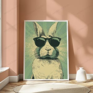 May include: A rectangular wall art print featuring a stylized illustration of a rabbit wearing black sunglasses. The rabbit is centered against a green background with a radiating sunburst pattern. The artwork uses a muted color palette.