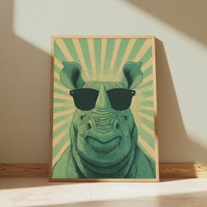 May include: A framed art print featuring a smiling rhinoceros wearing black sunglasses. The rhino is green, set against a retro-style background with green and tan rays. The artwork has a vintage, pop art aesthetic.