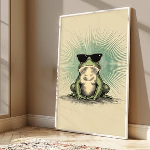 May include: A framed wall art print featuring a green frog wearing black sunglasses. The frog is centered against a light cream background with a teal sunburst graphic radiating from behind its head. The illustration uses detailed line work.