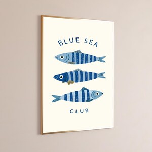 Pode incluir: Uma impress&atilde;o de arte de parede retangular num fundo creme com tr&ecirc;s peixes listrados azuis alinhados horizontalmente. O texto "BLUE SEA" est&aacute; impresso a azul acima dos peixes e a palavra "CLUB" est&aacute; impressa a azul abaixo.