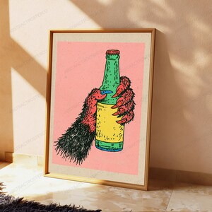 May include: A rectangular art print featuring a stylised illustration of a furry, clawed hand with black and red fur holding a green glass bottle with a yellow label. The background is a solid light pink colour, and the entire image is framed.