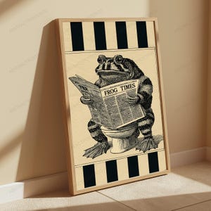 May include: A framed vertical poster featuring a black and white illustration of a toad sitting on a toilet while reading a newspaper titled THE FROG TIMES. The background consists of a cream-colored paper texture with black vertical stripes.