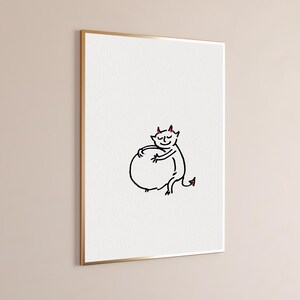 May include: A framed wall art print featuring a simple black line drawing of a small creature with red horns and a red-tipped tail, smiling while hugging a large round shape, set against a plain white background within a thin gold frame.