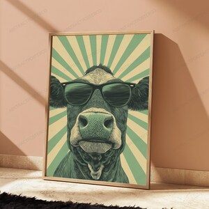 May include: A framed wall art print featuring a stylized illustration of a cow wearing black sunglasses. The cow is centered against a background of alternating light green and cream-colored sunburst rays. The artwork has a muted color palette.