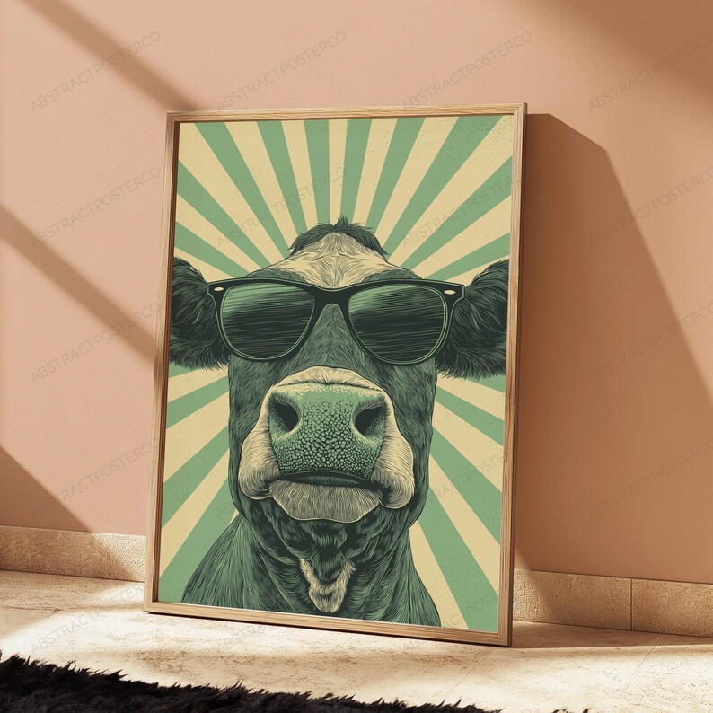 May include: A framed wall art print featuring a stylized illustration of a cow wearing black sunglasses. The cow is centered against a background of alternating light green and cream-colored sunburst rays. The artwork has a muted color palette.