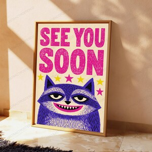 May include: A rectangular wall art print featuring a purple raccoon illustration with a wide grin. Above the raccoon, the text SEE YOU SOON is printed in bold, textured pink letters. A row of small yellow and pink stars sits between the text and the raccoon.