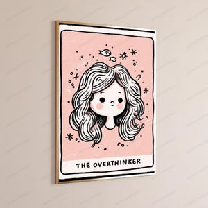 May include: A rectangular wall art print in a gold frame featuring a pink background with a black and white line drawing of a person with wavy hair. The text THE OVERTHINKER is printed at the bottom, surrounded by small star and fish illustrations.