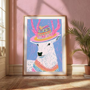 K&ouml;nnte beinhalten: Ein gerahmter Kunstdruck, der einen stilisierten wei&szlig;en Hirsch mit rosa Geweih zeigt, der einen farbenfroh gemusterten Hut mit breiter rosa Krempe und ein passendes gestreiftes Halstuch tr&auml;gt, vor einem leuchtend blauen Hintergrund.