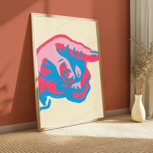 May include: A framed vertical wall art print featuring a stylised, abstract graphic of a bull head in vibrant shades of pink, light pink, and bright blue against a textured, off-white background. The artwork is enclosed in a thin gold frame.