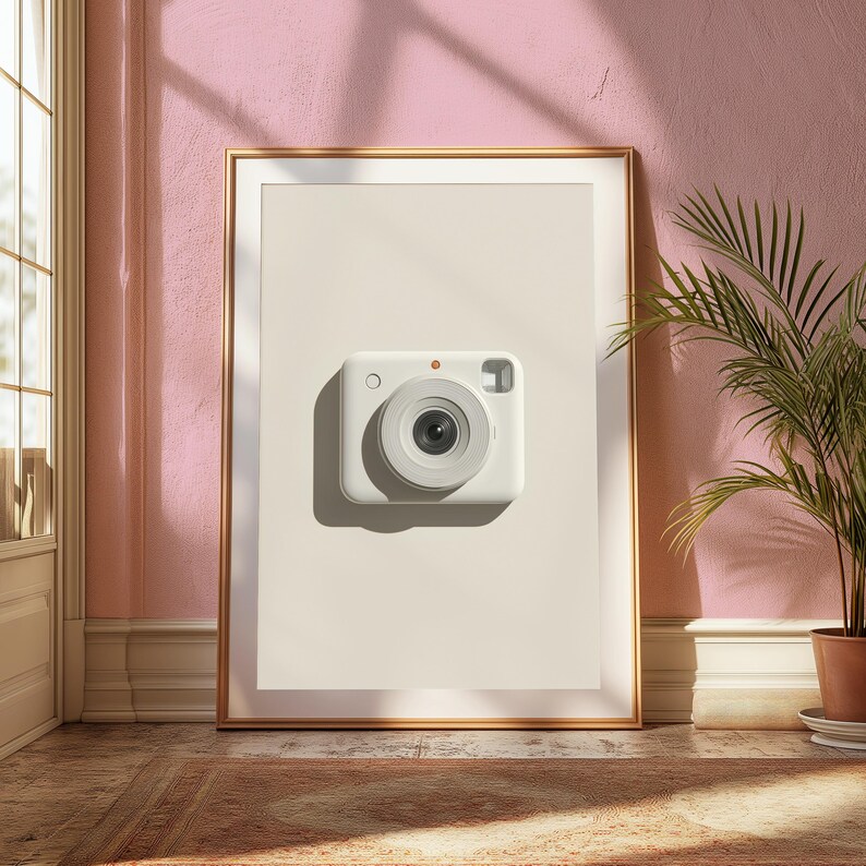 May include: A framed wall art print featuring a centered, minimalist white instant camera on a solid beige background. The rectangular frame is metallic gold, and the artwork displays a clean, modern composition with a single camera lens.