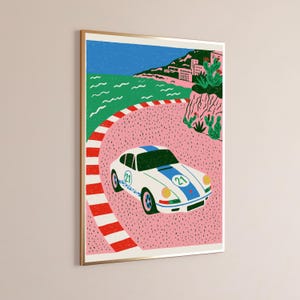 K&ouml;nnte beinhalten: Ein gerahmter Kunstdruck zeigt einen wei&szlig;en Sportwagen mit blauen Streifen und der Nummer 21 an der Seite, der auf einer rosa Rennstrecke mit rot-wei&szlig;er Begrenzung f&auml;hrt. Im Hintergrund sind ein gr&uuml;nes Meer, eine Felsk&uuml;ste und ein blauer Himmel.
