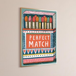 Op de afbeelding: Een ingelijste wandkunstprint met een gestileerde illustratie van een open luciferdoosje. Het doosje toont de tekst PERFECT MATCH in witte letters op een oranje achtergrond, omringd door kleurrijke patronen en een rij lucifers met gekleurde koppen.