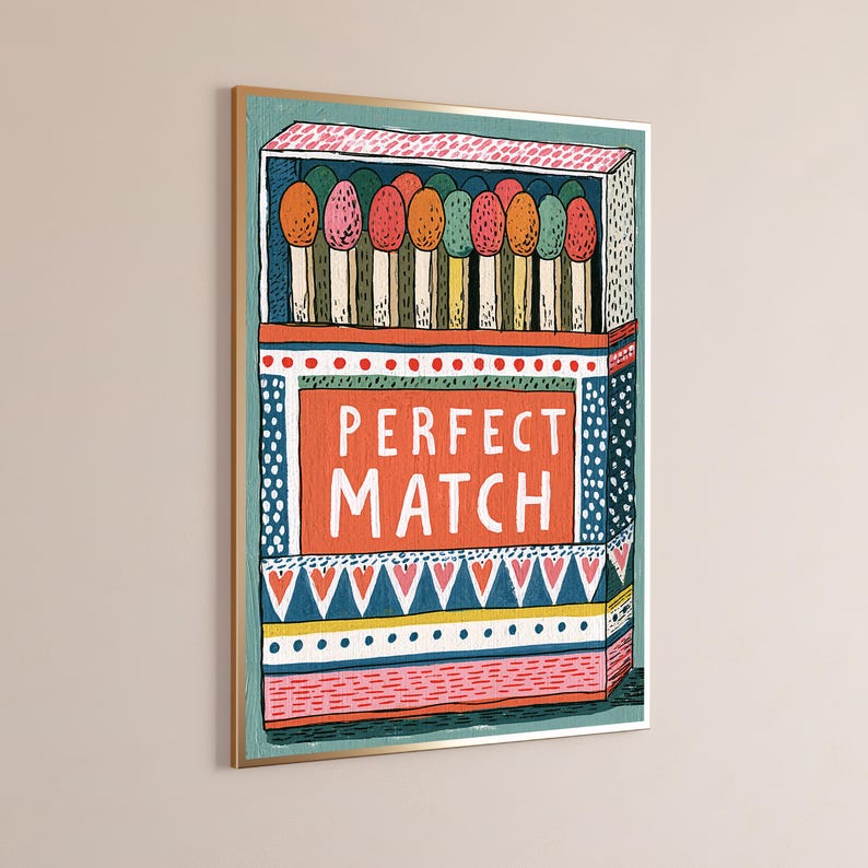 Op de afbeelding: Een ingelijste wandkunstprint met een gestileerde illustratie van een open luciferdoosje. Het doosje toont de tekst PERFECT MATCH in witte letters op een oranje achtergrond, omringd door kleurrijke patronen en een rij lucifers met gekleurde koppen.