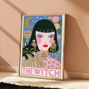 May include: A framed art print featuring a stylized illustration of a person with dark hair, a black cat, and pink floral patterns against a blue background. The text THE WITCH is printed in large pink capital letters at the bottom of the image.