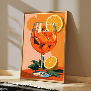 May include: A framed art print featuring a vibrant orange cocktail in a stemmed glass filled with ice cubes. An orange slice garnish rests on the rim, while another halved orange and green leaves sit at the base against a warm orange background.