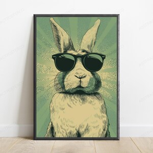 May include: A framed wall art print featuring a stylized illustration of a rabbit wearing black sunglasses. The rabbit is centered against a green background with a radiating sunburst pattern. The artwork uses a muted color palette with dark lines.