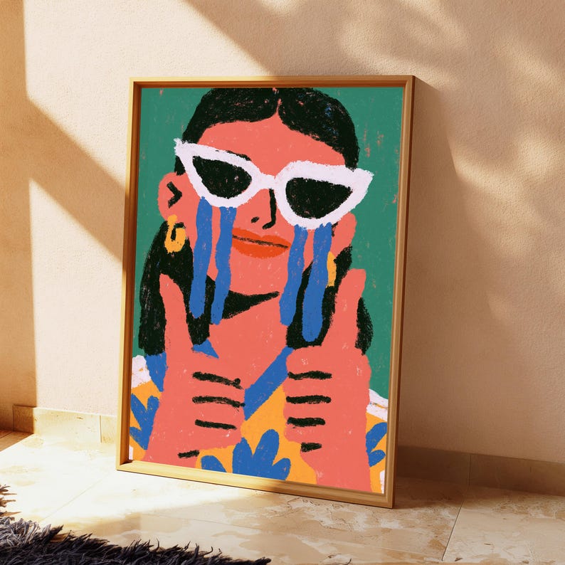 May include: A framed art print featuring a stylized illustration of a person with dark hair wearing white cat-eye sunglasses. The person has blue tears streaming down their face and is giving a double thumbs-up gesture against a green background.