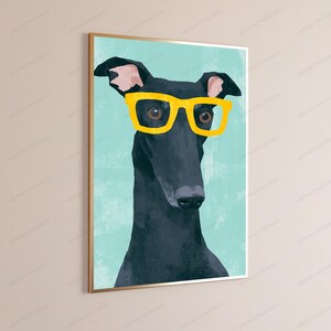 K&ouml;nnte beinhalten: Ein gerahmter Wandkunstdruck mit einer stilisierten Illustration eines schwarzen Windhundes, der eine leuchtend gelbe Brille tr&auml;gt. Der Hund ist vor einem einfarbigen hellblauen Hintergrund platziert und in einem d&uuml;nnen goldfarbenen Metallrahmen.