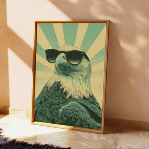 May include: A framed wall art print featuring a stylized illustration of a bald eagle wearing black sunglasses. The eagle is depicted against a background of alternating light green and beige sunburst rays with a dotted texture pattern.