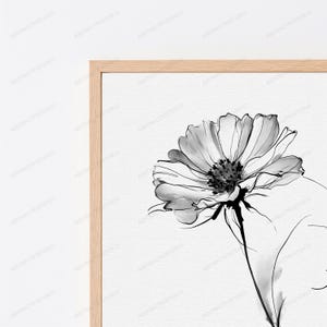 May include: A framed wall art print featuring a black and white ink wash illustration of a single flower with delicate petals and a dark centre. The artwork is encased in a light-toned wooden frame, showcasing a clean and simple aesthetic.