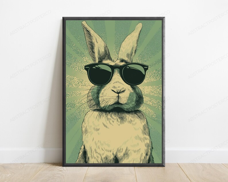 May include: A framed wall art print featuring a stylized illustration of a rabbit wearing black sunglasses. The rabbit is centered against a green background with a radiating sunburst pattern. The artwork uses a muted color palette with dark lines.
