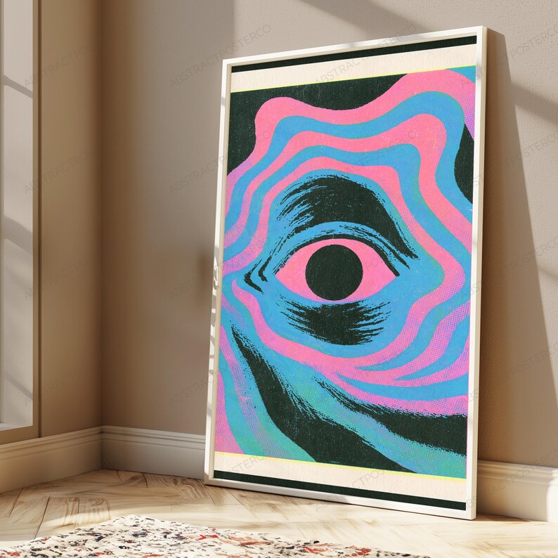 May include: A framed rectangular wall art print featuring a central black pupil surrounded by a pink iris and concentric wavy bands of alternating bright pink and light blue colors against a dark background, enclosed in a thin white frame.
