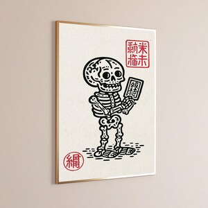 May include: A framed wall art print featuring a black ink illustration of a skeleton holding a small rectangular tablet. The design includes red stamp-like characters in the top right and bottom left corners on a textured off-white background.