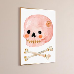 Puede incluir: Una impresi&oacute;n en acuarela enmarcada que muestra una calavera de color rosa claro con ojos circulares negros, nariz triangular negra y dientes dorados cosidos. La calavera tiene mejillas circulares naranjas y una peque&ntilde;a flor dorada. Dos huesos cruzados debajo.