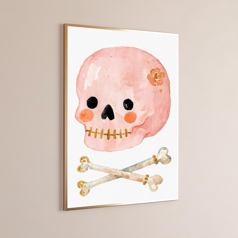 Puede incluir: Una impresi&oacute;n en acuarela enmarcada que muestra una calavera de color rosa claro con ojos circulares negros, nariz triangular negra y dientes dorados cosidos. La calavera tiene mejillas circulares naranjas y una peque&ntilde;a flor dorada. Dos huesos cruzados debajo.