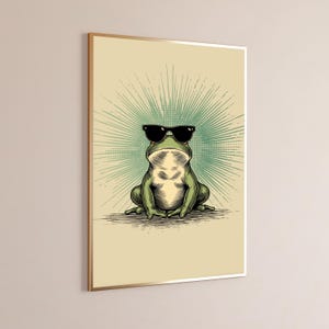 May include: A framed wall art print featuring a green frog wearing black sunglasses. The frog is centered against a light beige background with a radiating teal sunburst pattern behind it. The illustration uses bold lines and shading.
