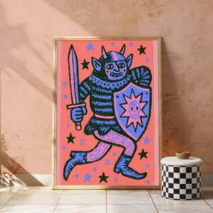 May include: A framed art print featuring a dark blue horned creature in armor holding a sword and a shield with a skull emblem. The background is a vibrant coral pink color decorated with scattered blue and black stars and small circles.