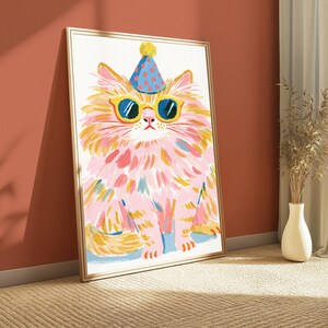 K&ouml;nnte beinhalten: Ein gerahmter Kunstdruck mit einem stilisierten Gem&auml;lde einer flauschigen Katze mit rosa und gelbem Fell. Die Katze tr&auml;gt einen blauen Partyhut mit roten Punkten und eine gelb umrandete Sonnenbrille mit dunkelblauen Gl&auml;sern auf wei&szlig;em Grund.