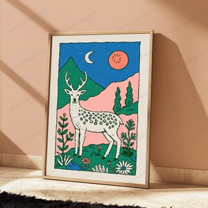May include: A rectangular wall art print in a light wood frame featuring a stylized illustration of a white spotted deer standing in a field of flowers. The background displays green mountains, a pink landscape, a blue night sky, a crescent moon, and a sun.